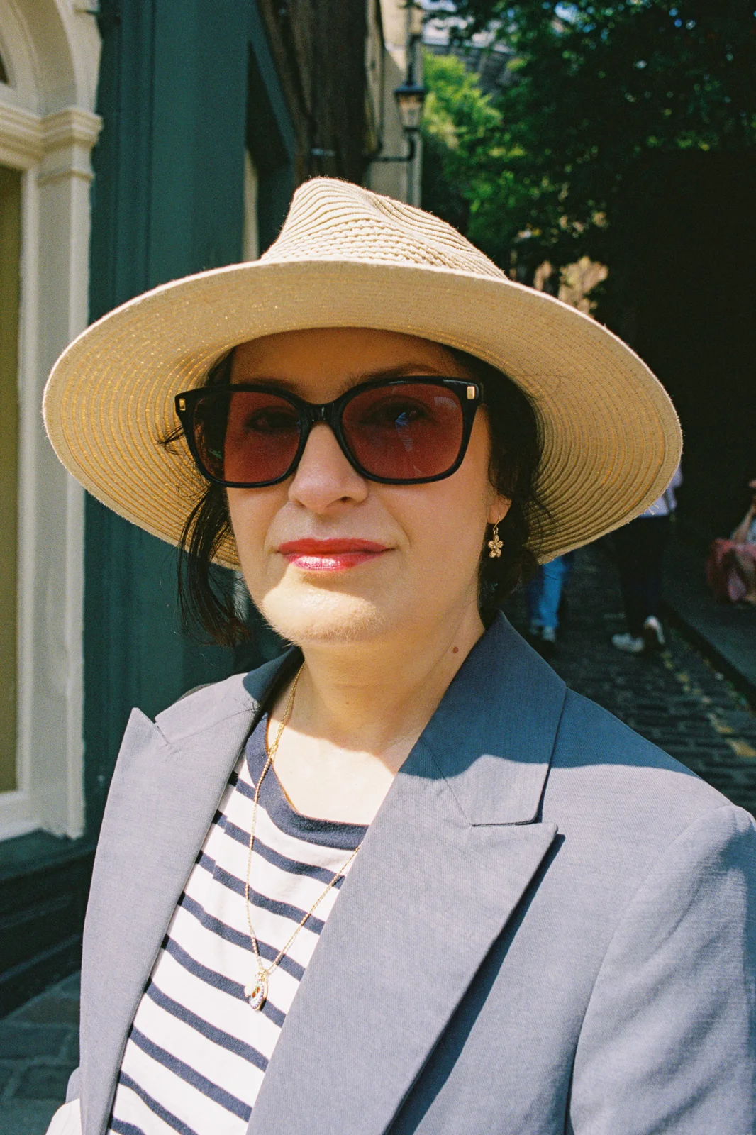 A woman in a straw hat, red-tinted sunglasses and a grey blazer on a cobbled street