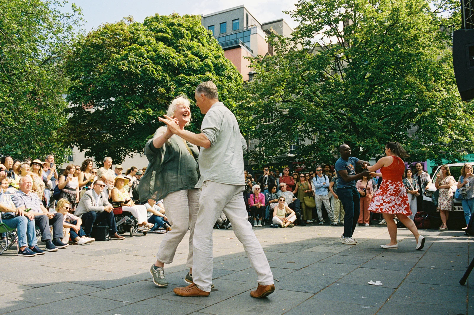 Two couples dancing in a city square in front of a seated crowd
