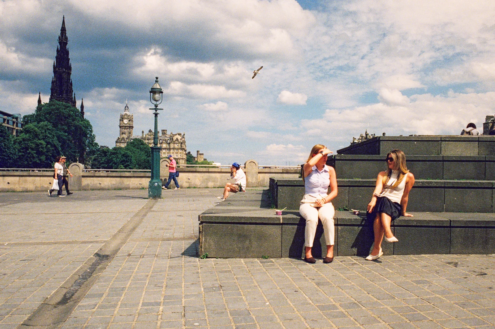 Two women sitting on stone steps in the sun, with the Scott Monument and Balmoral clock tower behind