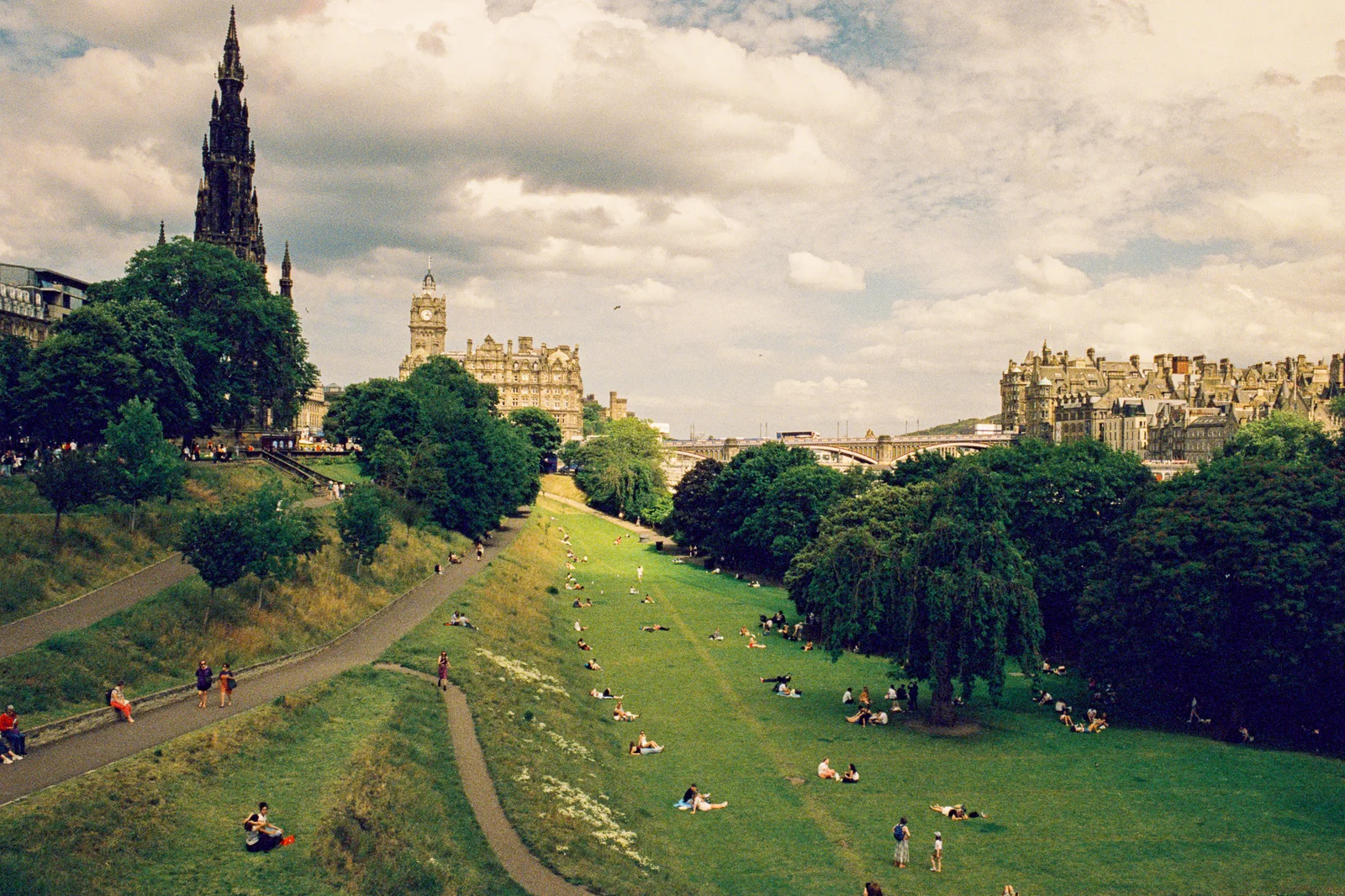 People lying on the grass in Princes Street Gardens under a cloudy sky, with the Scott Monument on the left