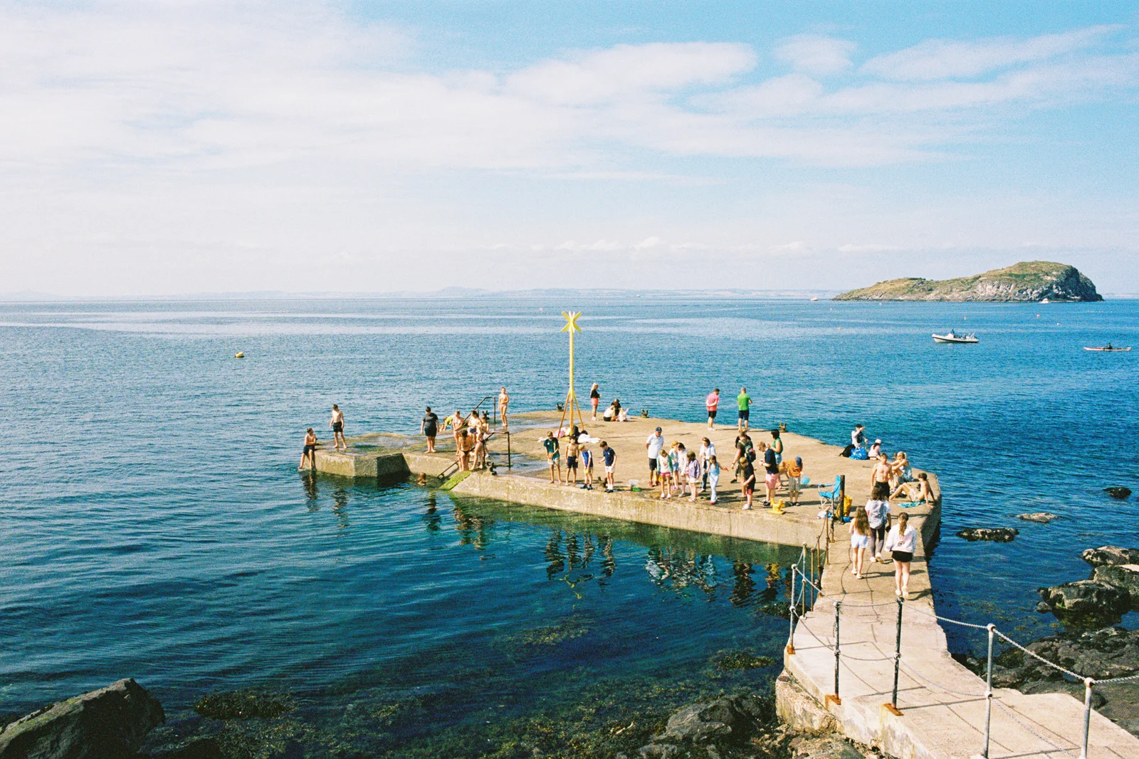 Swimmers and sunbathers on a concrete pier jutting into a calm blue sea, with an island on the horizon