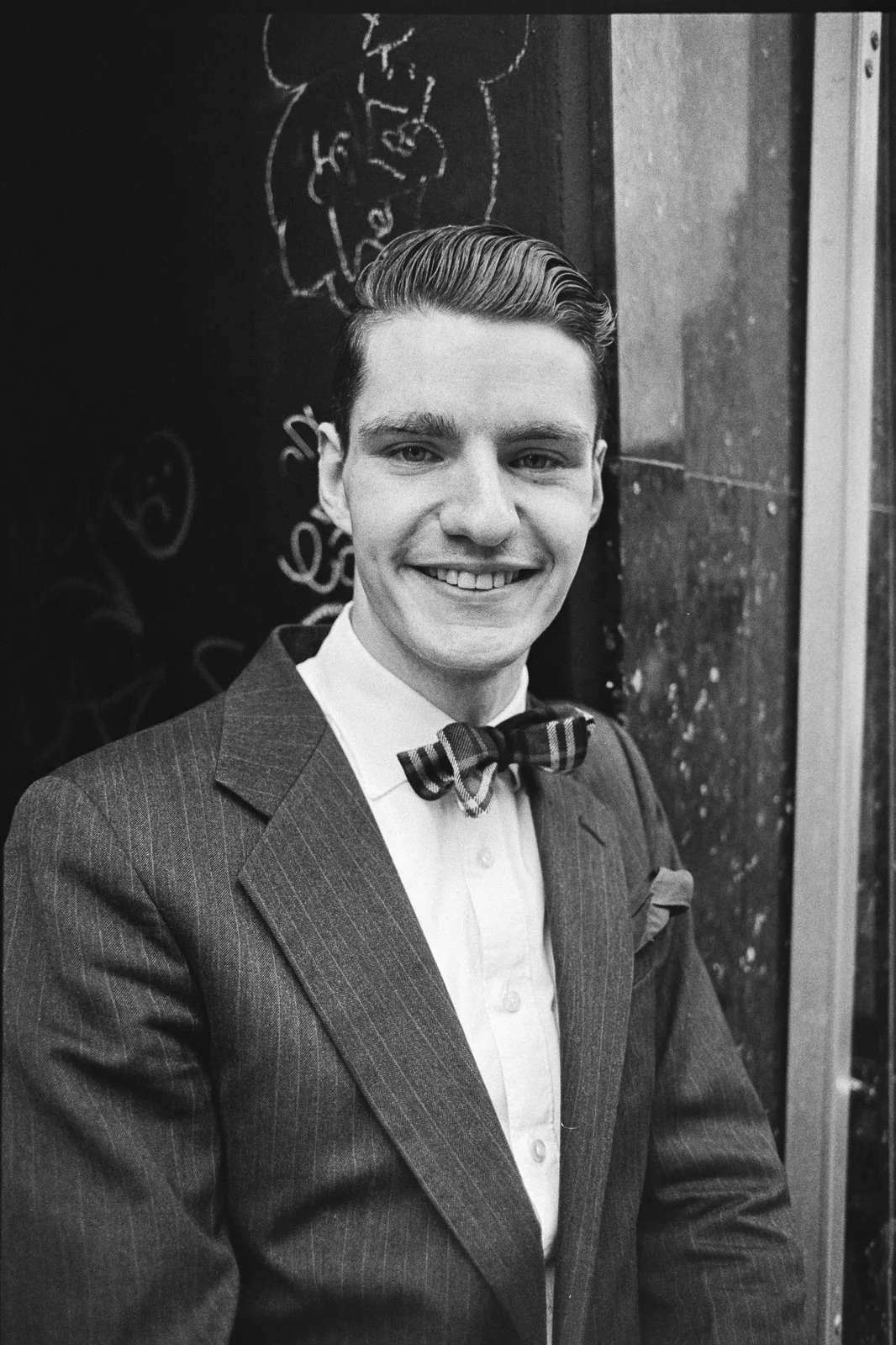 A smiling young man with slicked-back hair in a pinstripe suit and bow tie