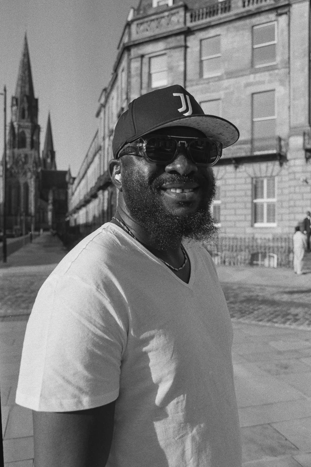 A smiling bearded man in a baseball cap and sunglasses on a sunny street